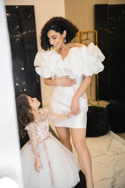 Happy and cute middle eastern little girl in floral attire looking at bride with wavy hair standing in trendy wedding dress with puff sleeves and ruffles in bridal boutique, mother and daughter — Stock Photo