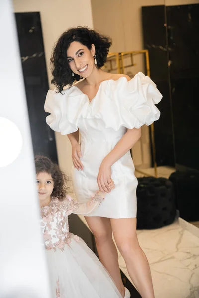 Happy middle eastern little girl in floral attire looking at mirror near bride with wavy hair standing in trendy wedding dress with puff sleeves and ruffles in bridal boutique, mother and daughter — Stock Photo