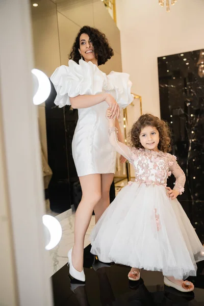 Счастливая ближневосточная невеста с волнистыми волосами, стоящая в модном свадебном платье с пышными рукавами и оборками, глядя на зеркало рядом с милой маленькой дочерью в цветочном наряде в свадебном бутике — стоковое фото