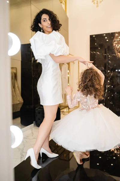Happy middle eastern bride with wavy hair in trendy wedding dress with puff sleeves and ruffles looking at mirror while dancing with cute little daughter in floral attire in bridal boutique — Stock Photo