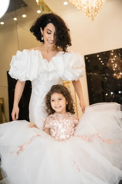 Joyful middle eastern little girl in floral attire holding tulle skirt and looking at mirror near bride with wavy hair standing in wedding dress with puff sleeves and ruffles in bridal boutique — Stock Photo