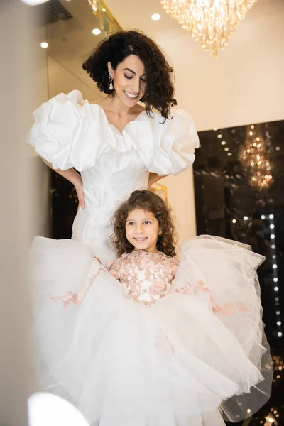 Joyful middle eastern little girl in floral attire holding tulle skirt and looking away near mother with wavy hair standing in white wedding dress with puff sleeves and ruffles in bridal boutique — Stock Photo