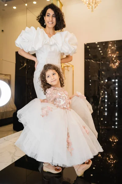 Charming middle eastern bride with brunette hair standing in white wedding gown with puff sleeves and ruffles and looking at mirror near daughter holding tulle skirt in bridal store — Stock Photo