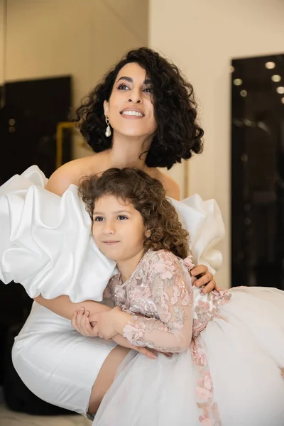 Charming middle eastern bride with brunette hair in white wedding dress with puff sleeves and ruffles hugging daughter and looking away in bridal store, special occasion — Stock Photo