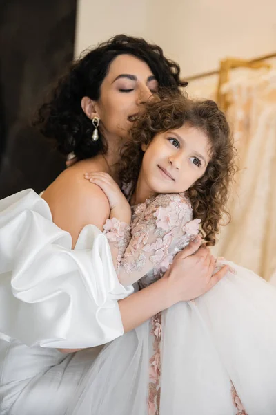 Enchanting middle eastern bride with brunette hair and closed eyes in white wedding dress with puff sleeves and ruffles hugging daughter looking away in bridal store, special occasion — Stock Photo