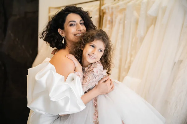 Charming middle eastern bride with brunette hair in white wedding dress with puff sleeves and ruffles embracing positive daughter in bridal store, white tulle fabrics on blurred background — Stock Photo