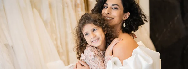 Gorgeous middle eastern bride with brunette hair in white wedding dress with puff sleeves and ruffles embracing positive daughter in bridal store, white tulle fabrics on blurred background, banner — Stock Photo