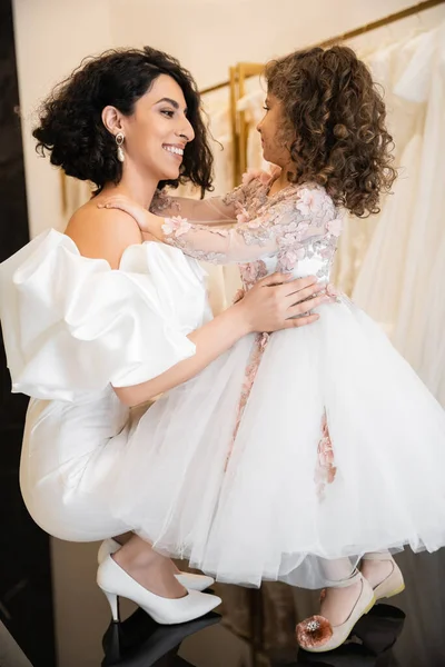 Happy middle eastern bride in white wedding dress with puff sleeves and ruffles embracing cheerful little daughter in bridal store, white tulle fabrics on blurred background, high heels — Stock Photo