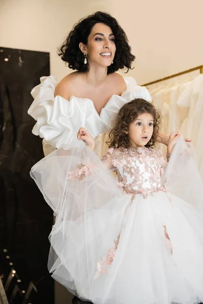 Captivating middle eastern bride with brunette hair standing in white wedding gown with puff sleeves and ruffles and looking away while holding tulle skirt of surprised daughter in bridal store — Stock Photo
