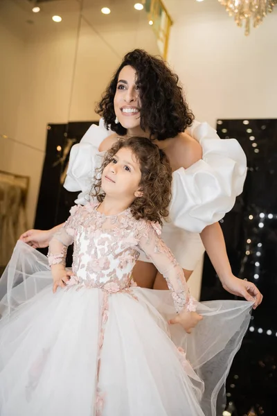 Charming middle eastern bride with brunette hair standing in white wedding gown with puff sleeves and ruffles and looking away while holding tulle skirt of daughter in bridal store — Stock Photo