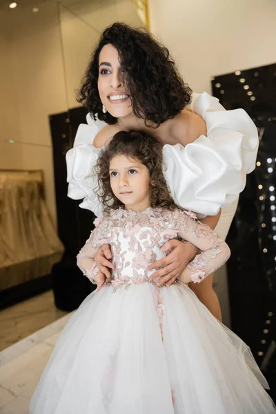Gorgeous middle eastern bride with brunette hair standing in white wedding dress with puff sleeves and ruffles behind cute daughter and smiling together in bridal store, looking away — Stock Photo