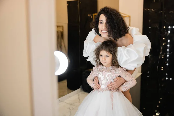 Charming middle eastern bride with brunette hair standing in white wedding gown with puff sleeves and ruffles and looking at mirror while hugging daughter with tulle skirt in bridal store — Stock Photo