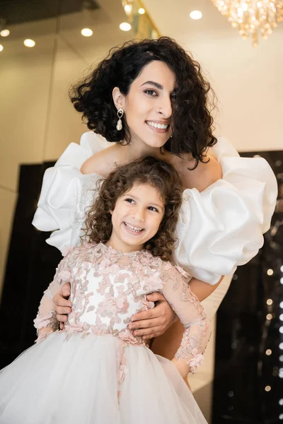 Gorgeous middle eastern bride with brunette hair standing in white wedding dress with puff sleeves and ruffles behind cute daughter and smiling together in bridal store, looking at camera — Stock Photo