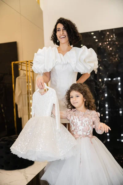 Charming middle eastern bride with brunette hair standing in white wedding gown with puff sleeves and ruffles and holding girly dress with tulle skirt near daughter in bridal store — Stock Photo
