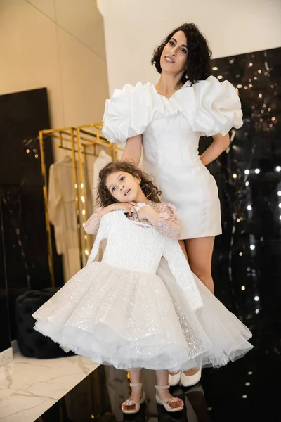 Charming middle eastern bride with brunette hair standing in white wedding gown with puff sleeves and ruffles near cute daughter holding girly dress with tulle skirt in bridal boutique — Stock Photo
