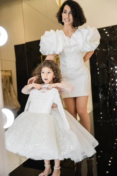 Happy middle eastern bride with brunette hair standing in white wedding gown with puff sleeves and ruffles near surprised daughter holding girly dress with tulle skirt near mirror in boutique — Stock Photo