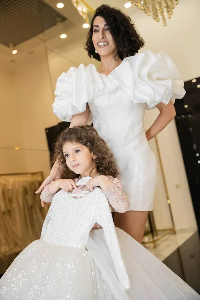Cheerful middle eastern woman with brunette hair standing in white wedding gown with puff sleeves and ruffles near cute daughter holding girly dress with tulle skirt in bridal boutique — Stock Photo