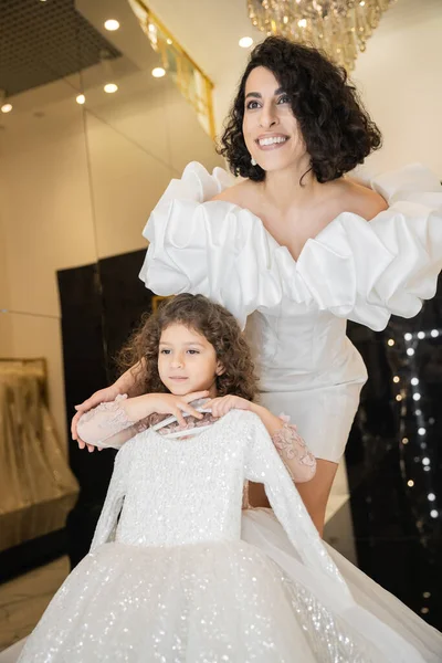 Joyous middle eastern bride with brunette hair standing in white wedding gown with puff sleeves and ruffles near cute daughter holding girly dress with tulle skirt in bridal boutique — Stock Photo