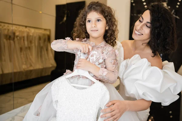 Charming middle eastern bride with brunette hair standing in white wedding gown with puff sleeves and ruffles looking at cute daughter holding girly dress with tulle skirt in bridal boutique — Stock Photo