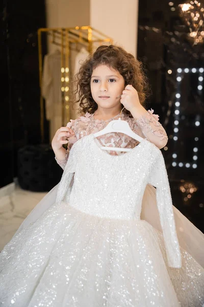 Curly middle eastern flower girl in attire holding hanger with white girly dress with tulle skirt in bridal boutique, preparation for wedding, blurred background, golden rack, choosing dress — Stock Photo