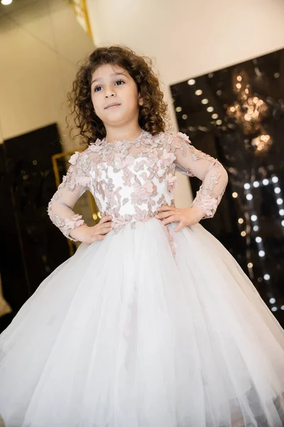 Low angle view of middle eastern little girl in floral dress with tulle skirt standing with hands on hips and looking away in bridal boutique, preparation for wedding, blurred background — Stock Photo
