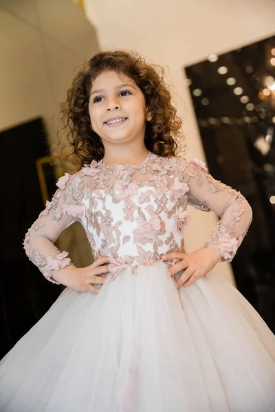 Cheerful middle eastern flower girl in floral dress with tulle skirt standing with hands on hips and looking away in bridal boutique, preparation for wedding, blurred background — Stock Photo