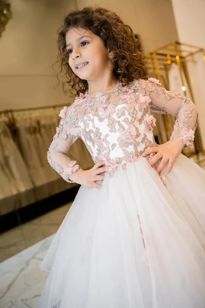 Радостный ближневосточный ребенок в цветочном платье с юбкой тюля, стоящий с руками на бедрах и глядя в свадебный бутик, подготовка к свадьбе, размытый фон, низкий угол, цветочница — стоковое фото
