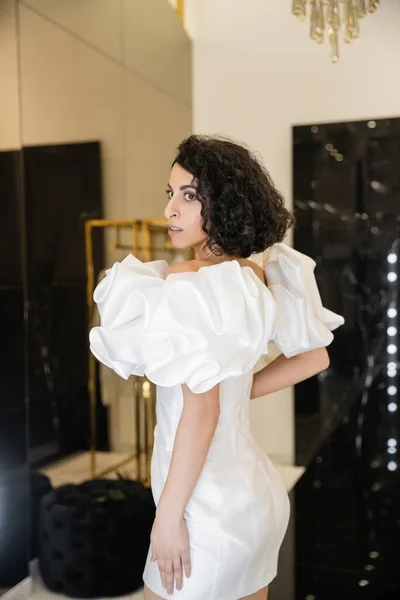 Middle eastern woman with brunette wavy hair trying on trendy wedding dress with puff sleeves and ruffles and looking away in modern bridal boutique, reflection, shopping, bride-to-be, exquisite — Stock Photo