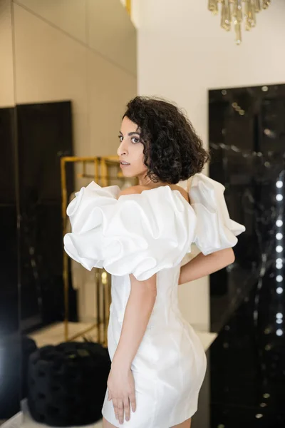 Femme du Moyen-Orient avec des cheveux bruns ondulés essayant sur la robe de mariée à la mode avec des manches bouffantes et des volants et regardant loin dans la boutique de mariée moderne, réflexion, shopping, future mariée, exquis — Photo de stock