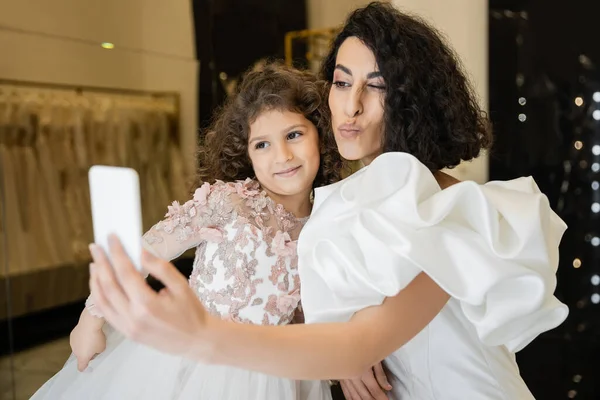 Noiva do Oriente Médio com cabelo morena em vestido de noiva branco com mangas de sopro e babados tomando selfie no smartphone enquanto amuada lábios perto da filha feliz na loja de noivas — Fotografia de Stock
