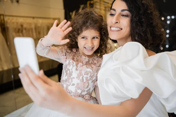 Charmante mariée du Moyen-Orient avec des cheveux bruns en robe de mariée blanche avec des manches bouffantes et des volants tenant smartphone tandis que la fille heureuse agitant la main pendant l'appel vidéo dans le magasin de mariée — Photo de stock