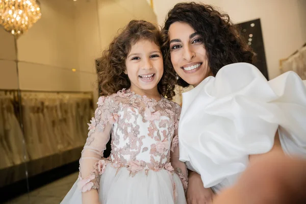Fröhliches kleines Mädchen in floraler Kleidung lächelt neben charmanter Mutter im weißen Hochzeitskleid mit Puffärmeln und Rüschen, während sie gemeinsam in die Kamera in der Brautboutique guckt, Selfie — Stockfoto