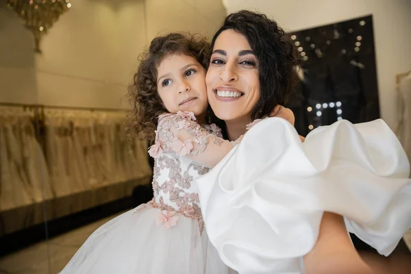 Cheerful little girl in floral attire hugging tight her charming mother in white wedding dress with puff sleeves and ruffles while smiling and looking at camera in bridal boutique — Stock Photo