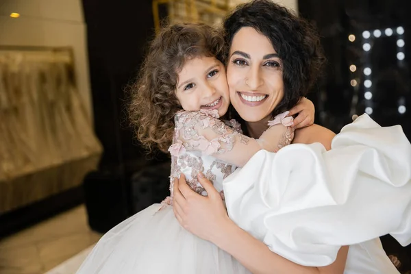 Glückliches kleines Mädchen in floraler Kleidung umarmt ihre charmante Mutter im weißen Hochzeitskleid mit Puffärmeln und Rüschen, während sie lächelt und gemeinsam in die Kamera in der Brautboutique schaut — Stockfoto