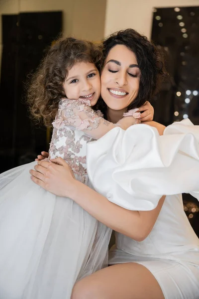 Fröhliches kleines Mädchen in floraler Kleidung umarmt ihre charmante Mutter aus dem Nahen Osten mit geschlossenen Augen im weißen Hochzeitskleid mit Puffärmeln und Rüschen, während sie in der Brautboutique lächelt — Stockfoto