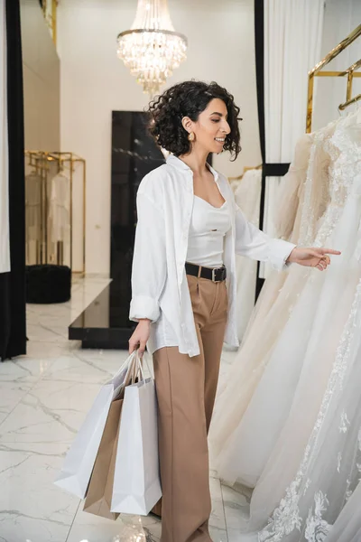 Stylische und glückliche Frau aus dem Mittleren Osten mit brünetten und welligen Haaren, die in einer beigen Hose mit weißem Hemd steht und Einkaufstaschen hält, während sie im Brautsalon das Brautkleid auswählt — Stockfoto
