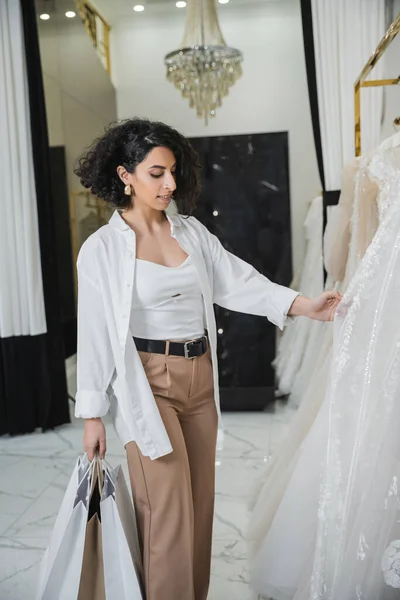 Encantadora mulher do Oriente Médio com morena e cabelo ondulado de pé em calças bege com camisa branca e segurando sacos de compras ao escolher o vestido de noiva no salão de casamento, shopping spree — Fotografia de Stock