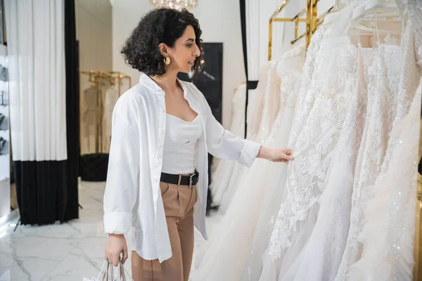 Side view of happy middle eastern woman with brunette and wavy hair standing in beige pants with white shirt and holding shopping bags while choosing wedding dress in bridal boutique — Stock Photo
