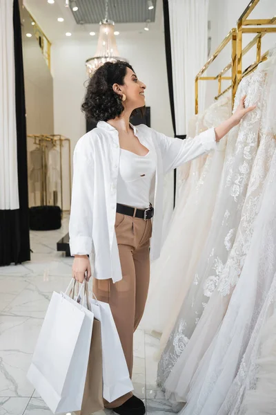 Стильная и радостная средневосточная женщина с брюнеткой и волнистыми волосами, стоящая в бежевых брюках в белой рубашке и держащая сумки, выбирая свадебное платье в свадебном бутике, шик — стоковое фото