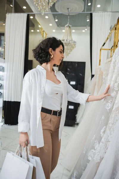 Side view of stylish and happy middle eastern woman with brunette and wavy hair standing in beige pants with white shirt and holding shopping bags while choosing wedding dress in bridal salon — Stock Photo