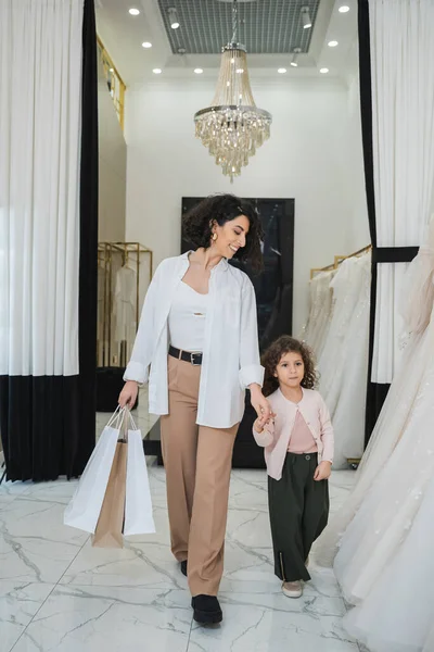 Glückliche Frau aus dem Nahen Osten mit brünetten Haaren in beiger Hose und weißem Hemd mit Einkaufstaschen beim Spaziergang mit kleiner Tochter in der Nähe von Brautkleidern im Brautsalon, moderne Braut, Mutter — Stockfoto