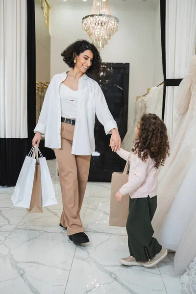 Happy middle eastern woman with brunette hair in beige pants with white shirt holding shopping bags and hand of little daughter near wedding dresses in bridal salon, modern bride, mother — Stock Photo
