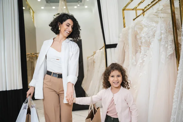 Mulher feliz do Oriente Médio com cabelos morena segurando sacos de compras enquanto caminhava com menina alegre perto de vestidos de noiva no salão de casamento, noiva moderna, mãe e filha, vínculo especial — Fotografia de Stock