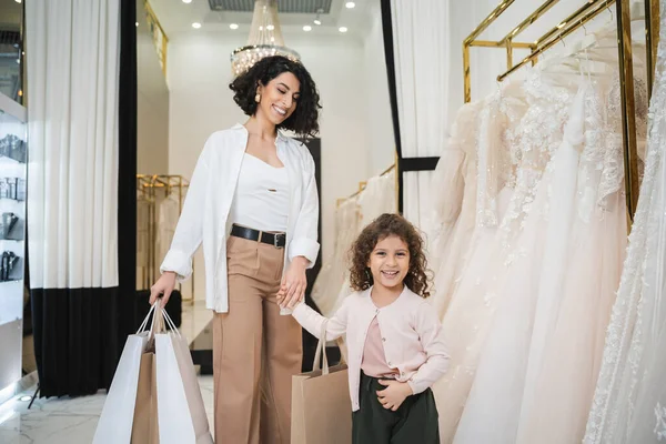 Happy middle eastern bride with brunette hair in beige pants with white shirt holding shopping bags while standing with little girl near wedding dresses in bridal salon, mother and daughter — Stock Photo
