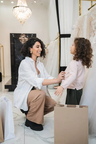 Cheerful middle eastern woman with brunette hair in white shirt sitting near shopping bags and wearing cardigan on little girl near wedding dresses in bridal salon, mother and daughter, bride — Stock Photo