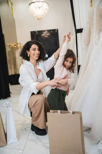 Cheerful middle eastern woman with brunette hair in white shirt sitting near shopping bags and holding hands of little girl dancing near wedding dresses in bridal salon, mother and daughter, bride — Stock Photo