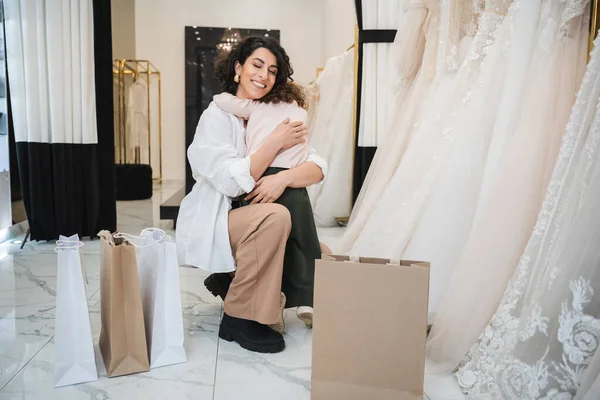 Little girl hugging tight cheerful middle eastern bride with brunette hair in white shirt sitting near shopping bags and wedding dresses in bridal salon, mother and daughter, bonding — Stock Photo