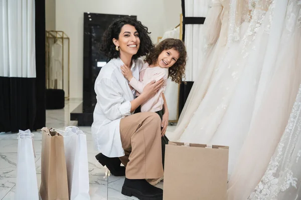 Счастливая маленькая девочка обнимает веселую невесту с брюнетками в белой рубашке, сидя рядом с пакетами покупок и свадебными платьями в свадебном салоне, мать и дочь, сближаясь — стоковое фото