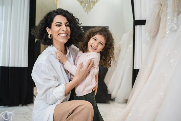 Happy little girl hugging cheerful middle eastern bride with brunette hair in white shirt sitting near white wedding dresses in bridal salon, mother and daughter, bonding, special moment, shopping — Stock Photo