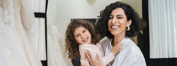Curly little girl hugging cheerful middle eastern bride with brunette hair in white shirt sitting near white wedding dresses in bridal salon, mother and daughter, bonding, special moment, banner — Stock Photo